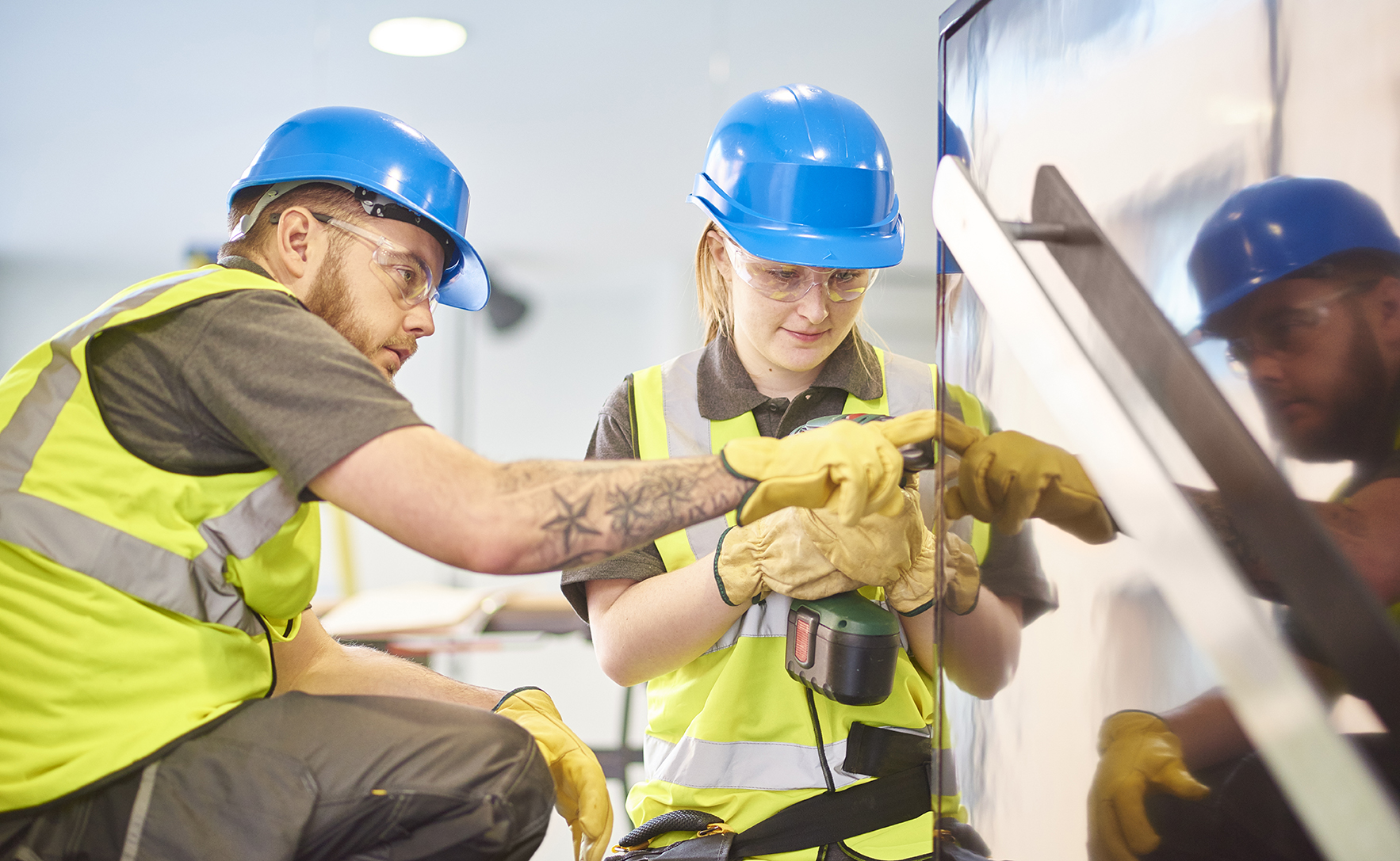 a teenage female apprentice carpenter is installing handrails  in a modern office refurbishment. She is being supervised by her co-worker . She is using a drill to make guide holes for the hand rails  . They are wearing safety workwear.